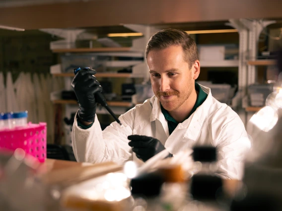 Zach Compton adds a solution to a tube using a pipette. He is wearing black gloves and has a white lab coat.